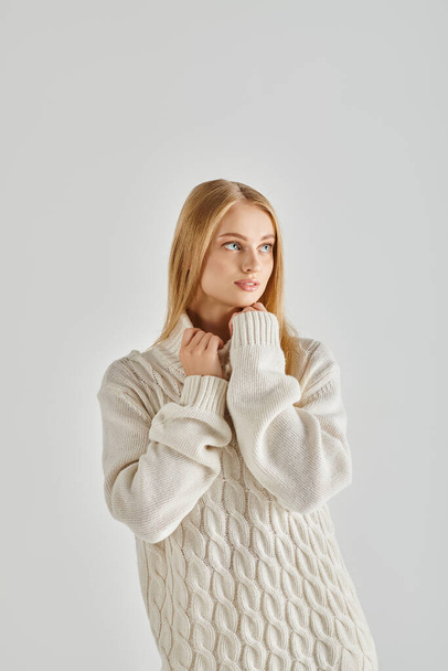 στοχαστική ξανθιά γυναίκα σε λευκό ζεστό πουλόβερ κοιτάζοντας κάμερα σε γκρι, συναισθήματα χειμώνα - Φωτογραφία, εικόνα