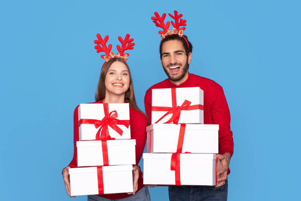 Χαρούμενος άνδρας και γυναίκα φορώντας τάρανδοι κόμμα κέρατα κέρατα κρατώντας στοίβα από κουτιά δώρων Χριστούγεννα, ευτυχισμένο ζευγάρι μεταφέρουν δώρα και χαμογελώντας στην κάμερα, ποζάρει απομονωμένη στο μπλε φόντο, αντίγραφο χώρο - Φωτογραφία, εικόνα