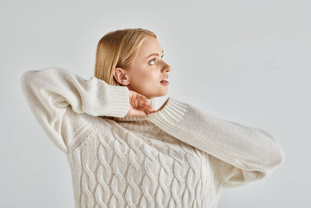 atrakcyjna i marzycielska kobieta w ciepłym swetrze stojąca i odwracająca wzrok na szarym, zimowym stylu - Zdjęcie, obraz