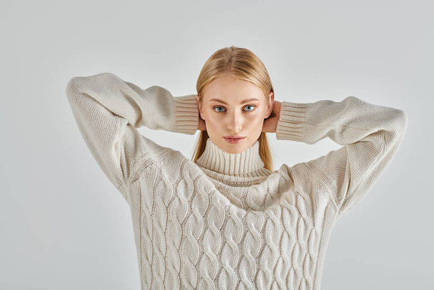 暖かいセーターの魅力的なブロンドの女性は,頭の後ろに手を保持し,灰色のカメラを見ています - 写真・画像