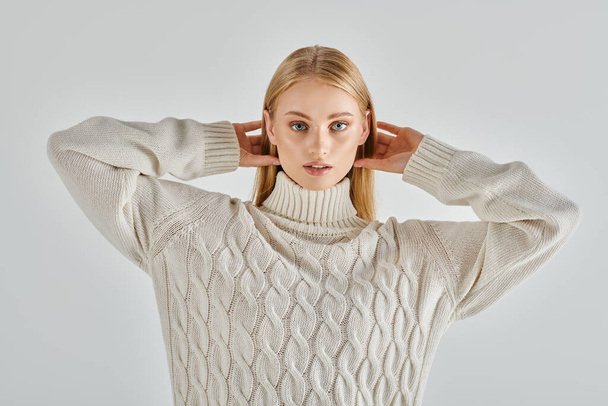 νεαρή κομψή γυναίκα με εκφραστικό βλέμμα και ξανθά μαλλιά ποζάρουν σε λευκό πλεκτό πουλόβερ σε γκρι - Φωτογραφία, εικόνα