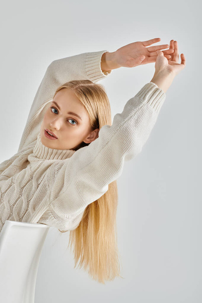молодая блондинка в вязаном свитере сидит и вытягивает руки, глядя в камеру на сером - Фото, изображение