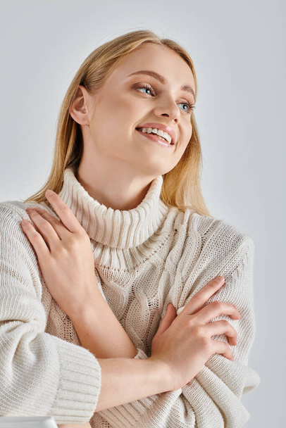carefree woman with blonde hair sitting in cozy winter sweater and looking away on grey, happiness - Photo, Image
