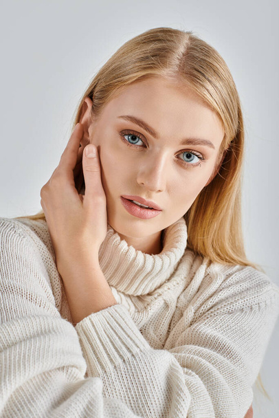 portrét smyslné blondýny v přírodním make-upu a bílém zimním svetru dotýkajícím se tváře na šedé - Fotografie, Obrázek