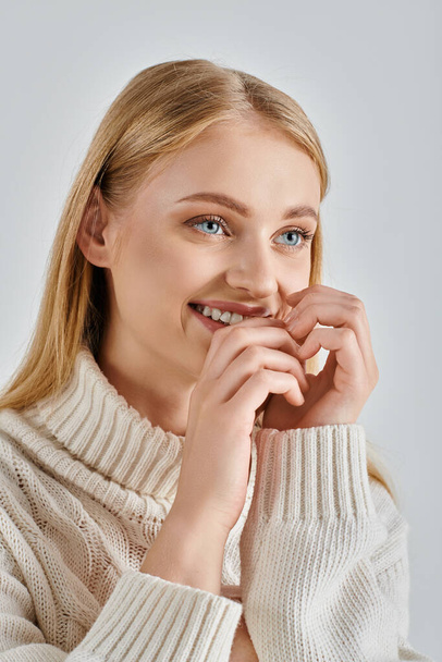 jeune femme joyeuse en pull blanc chaud et maquillage naturel souriant avec les mains près du visage sur gris - Photo, image