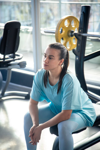 Уставшая молодая женщина в синей спортивной одежде отдыхает, сидя после тренировки в спортзале. - Фото, изображение