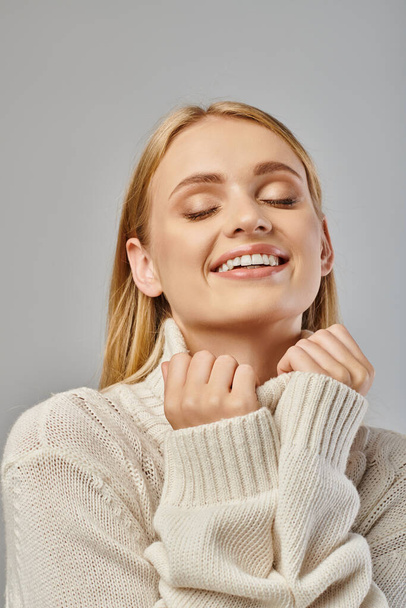delighted blonde woman in warm knitted sweater smiling with closed eyes on grey, winter fashion - Photo, Image