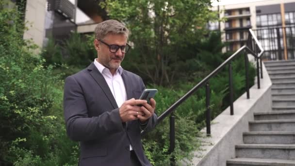 Усміхнений дорослий бізнесмен тримає телефон, позуючи біля сходів у місті, друкуючи. Бізнес, комунікація, концепція технології. Повільний рух - Кадри, відео