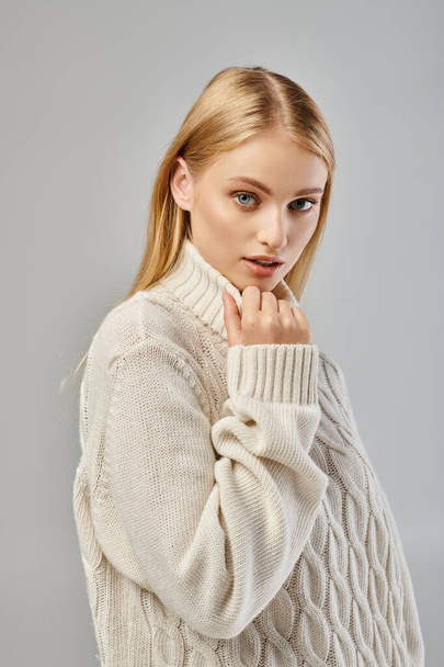 alluring woman with blonde hair and natural makeup wearing white sweater looking at camera on grey - Φωτογραφία, εικόνα