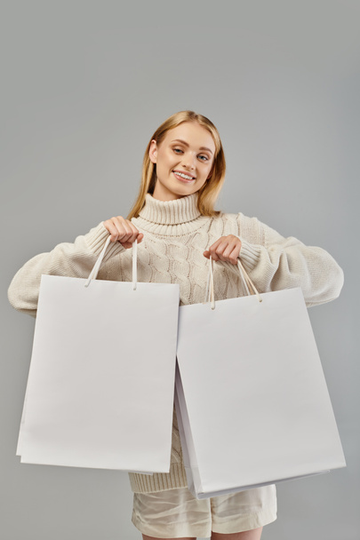 joyful blonde woman in white knitted  sweater showing shopping bags on grey, seasonal sales - Photo, Image