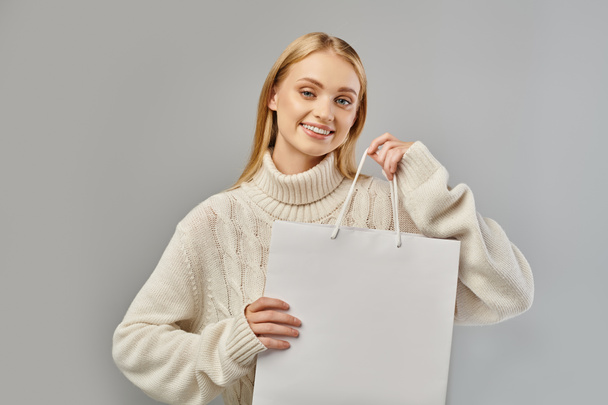 mujer rubia alegre en suéter de punto cálido con bolsa de compras blanca en gris, ventas de invierno - Foto, imagen