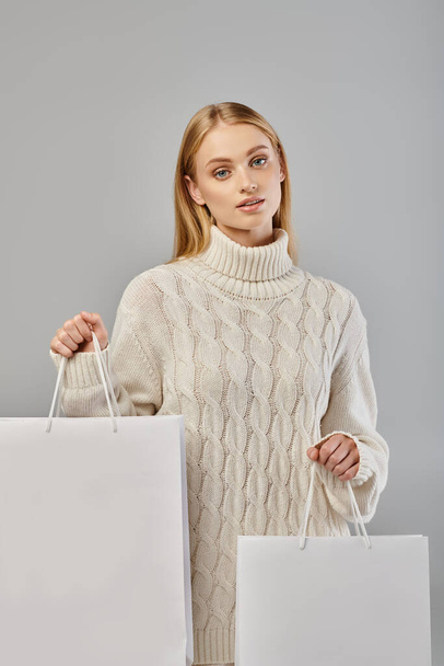 encantadora mujer rubia en suave suéter caliente de pie con bolsas de compras blancas en gris, las ventas de invierno - Foto, Imagen