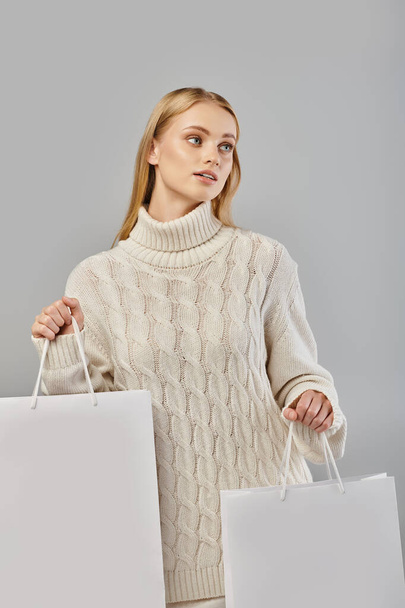 betoverende blonde vrouw in gezellige gebreide trui met witte boodschappentassen weg te kijken op grijs - Foto, afbeelding
