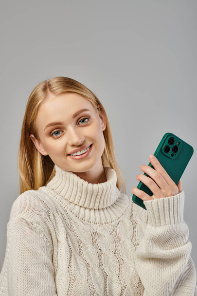 mujer rubia joven y alegre en suéter de invierno sosteniendo teléfono inteligente y mirando a la cámara en gris - Foto, imagen