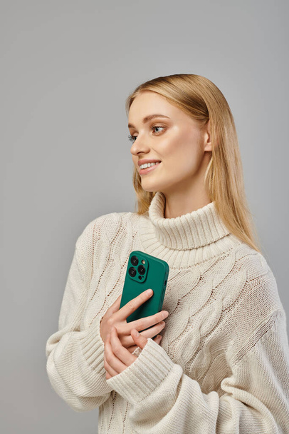 smiling and dreamy woman in delicate knitted sweater holding  smartphone and looking away on grey - Photo, Image
