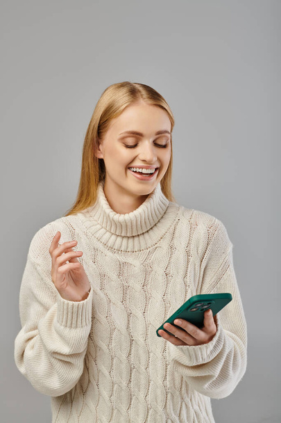 cheerful blonde woman in warm knitted sweater smiling during video call on smartphone on grey - Photo, Image