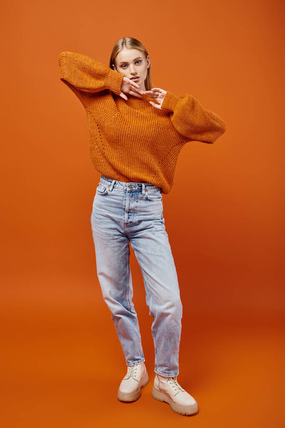 young blonde woman in bright knitted sweater and blue jeans posing on orange backdrop, full length - Photo, Image