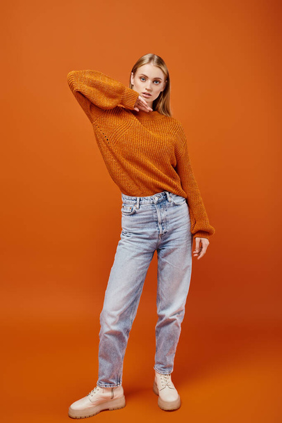 trendige blonde Frau in warmem Strickpullover und blauen Jeans vor orangefarbenem Hintergrund, Wintermode - Foto, Bild