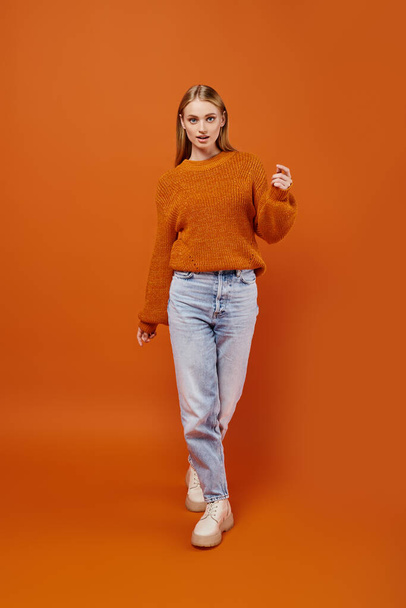 trendy blonde woman in blue jeans and orange winter sweater walking on bright backdrop, full length - Photo, Image