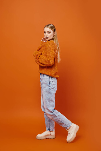 smyslná blondýna žena v modrých džínách a útulný zimní svetr při pohledu na kameru na oranžové pozadí - Fotografie, Obrázek