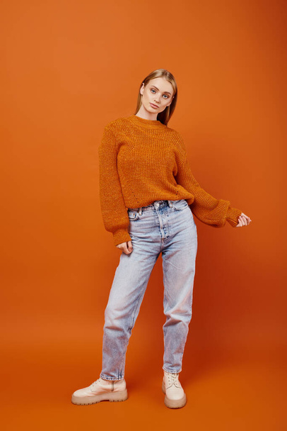 fashionable blonde woman in jeans and orange knitted sweater posing on bright backdrop, winter vibes - Photo, Image
