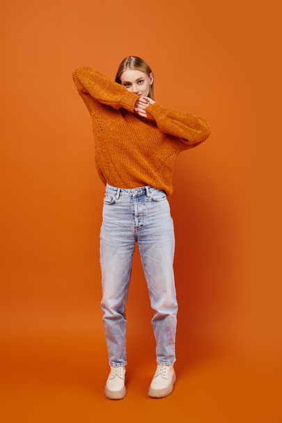 стильная блондинка в джинсах и оранжевом трикотаже смотрит в камеру на ярком фоне - Фото, изображение