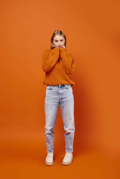 joyful blonde woman in jeans and warm knitted sweater smiling and looking away on orange backdrop - Photo, Image