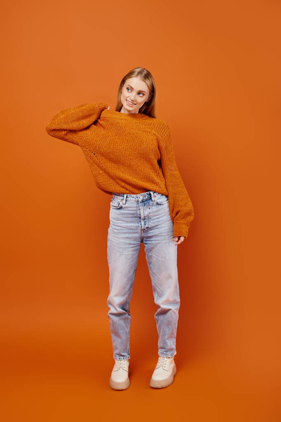 full length of trendy blonde woman in jeans and soft knitted sweater posing on orange backdrop - Photo, Image