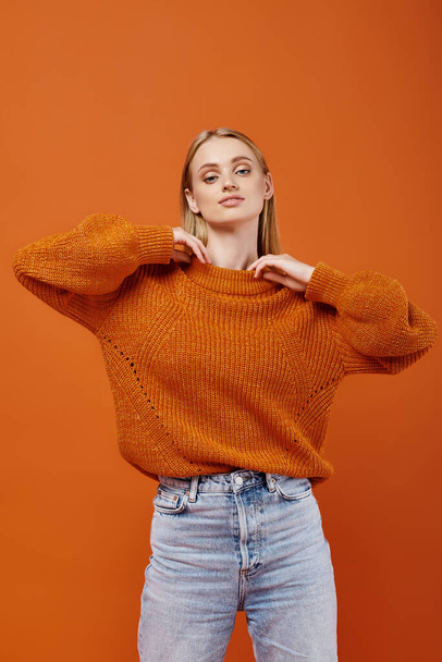 stylová blondýnka v teplém a jasném pleteném svetru a modrých džínách na oranžovém pozadí, móda - Fotografie, Obrázek