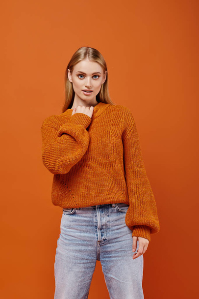 alluring blonde woman in bright knitted sweater and blue jeans looking at camera on orange backdrop - Photo, Image