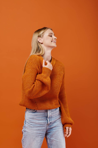 возбужденная блондинка в ярком свитере и синих джинсах смеется и смотрит в сторону на оранжевом фоне - Фото, изображение