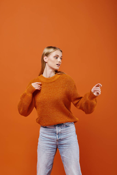 sensual blonde woman in bright sweater and blue jeans looking away on orange backdrop, winter style - Photo, Image