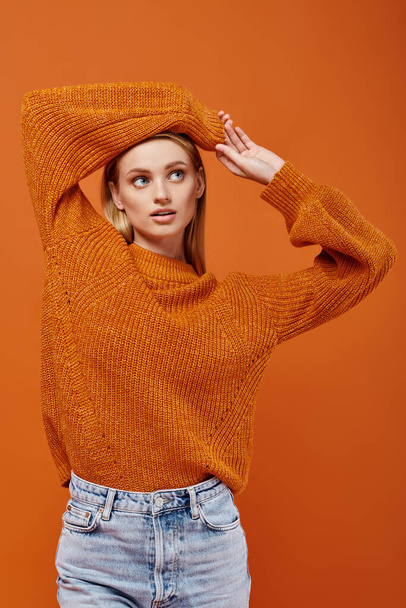 mladá blondýnka v oranžovém pleteném svetru pózuje s pažemi nad hlavou na oranžové, zimní vibrace - Fotografie, Obrázek