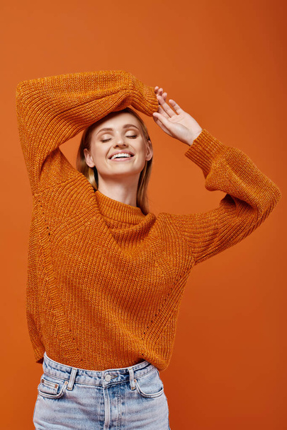 happy blonde woman in bright winter sweater with arms above head smiling with closed eyes on orange - Photo, Image