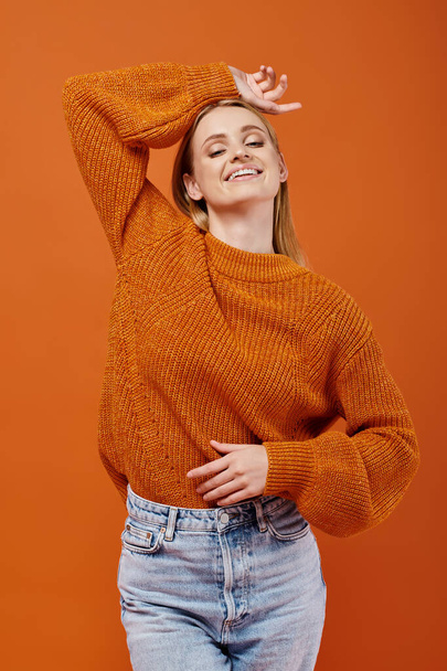 szczęśliwa blondynka w jasnym zimowym swetrze pozowanie z ręką nad głową na pomarańczowy, radość zima - Zdjęcie, obraz