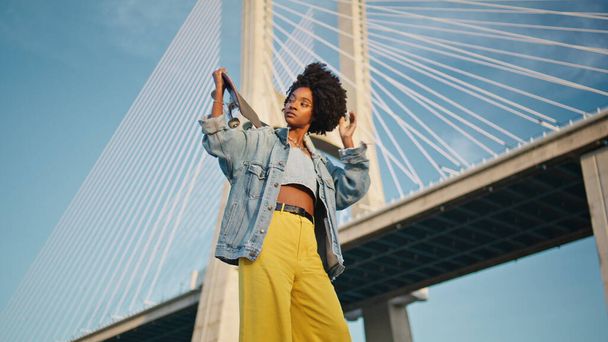 Piękna dziewczyna z deskorolką pozowanie przed mostem miejskim letni dzień. Fajny afrykański nastolatek trzymający longboard stojący na widoku błękitnego nieba. Kręcone modne nastolatek hipster patrząc na odległość - Zdjęcie, obraz