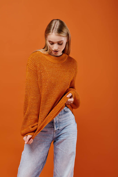 young blonde woman in blue jeans stretching her bright warm sweater on orange backdrop, fashion - Photo, Image