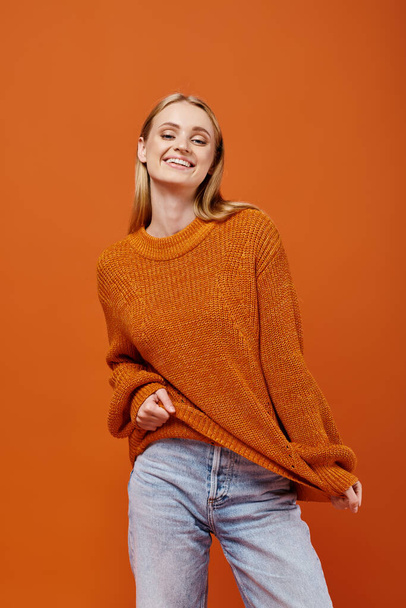 χαρούμενη ξανθιά γυναίκα τεντώνει το φωτεινό πουλόβερ της και χαμογελά στην κάμερα σε πορτοκαλί, χειμωνιάτικα συναισθήματα - Φωτογραφία, εικόνα