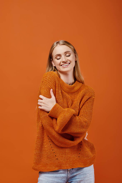 pleased woman in warm knitted sweater hugging herself and smiling with closed eyes on orange - Photo, Image