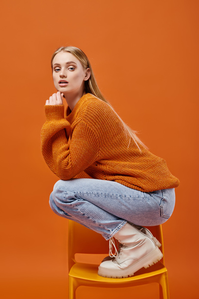 expressieve vrouw in gebreide wintertrui en jeans poserend op stoel en kijkend naar camera op oranje - Foto, afbeelding