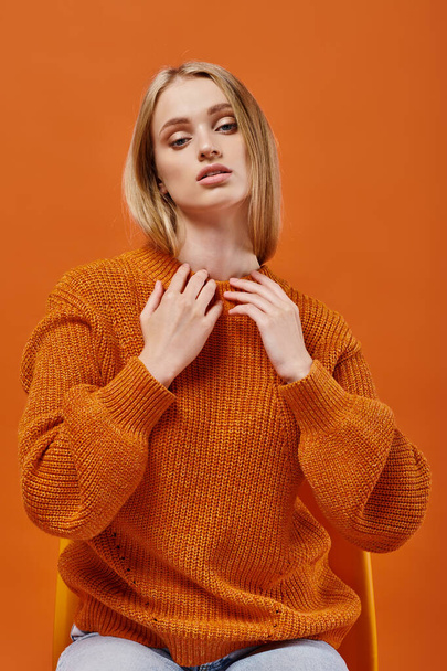 expressive blonde woman in bright winter sweater sitting on chair while looking at camera on orange - Photo, Image