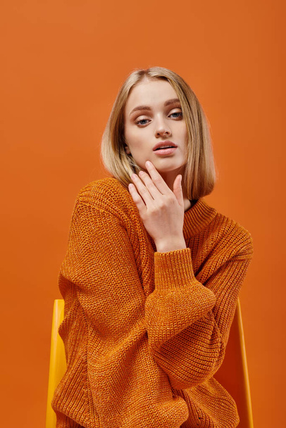 charming blonde woman in orange sweater sitting on chair and looking at camera on colorful backdrop - Fotoğraf, Görsel