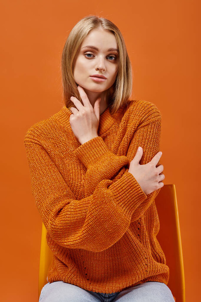 fashionable blonde woman in orange sweater sitting on chair and looking at camera on bright backdrop - Photo, Image