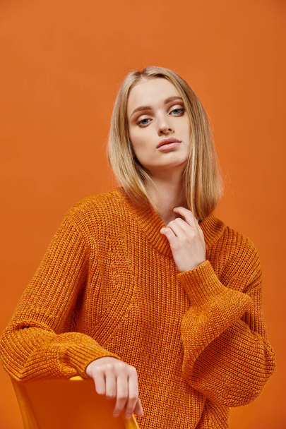 sophisticated blonde woman in orange sweater sitting and looking at camera on colorful backdrop - Photo, Image