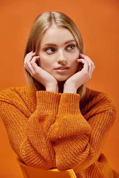 dromerige blonde vrouw in lichte en zachte gebreide trui met natuurlijke make-up weg te kijken op oranje - Foto, afbeelding
