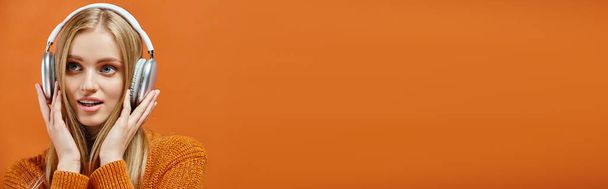 Ilık örülü süveter giymiş, turuncu, yatay afişle kulaklıkla müzik dinleyen ilham verici bir kadın. - Fotoğraf, Görsel
