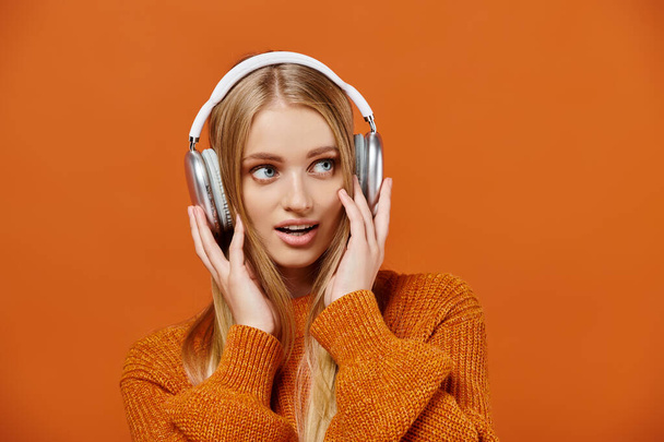 mujer rubia sorprendida en suéter de punto brillante escuchando música en auriculares en naranja, disfrute - Foto, imagen