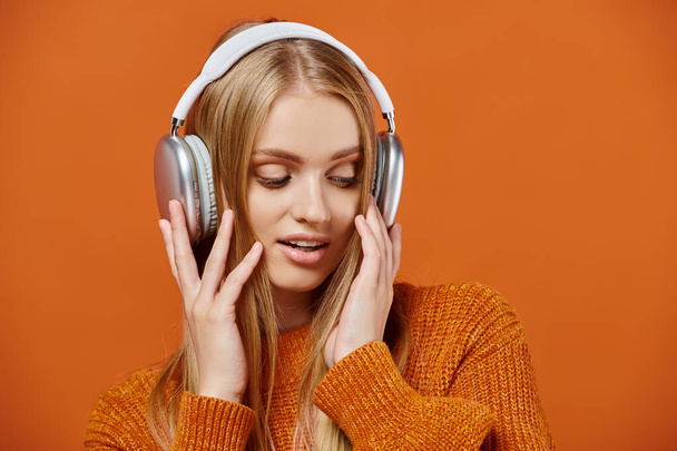 オレンジ色の背景で音楽を聞いているカラフルな編まれたセーターとヘッドフォンの魅力的なブロンドの女性 - 写真・画像