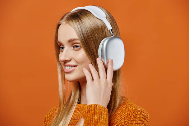 портрет улыбающейся блондинки в тёплом ярком свитере и наушниках, слушающей музыку на оранжевом - Фото, изображение