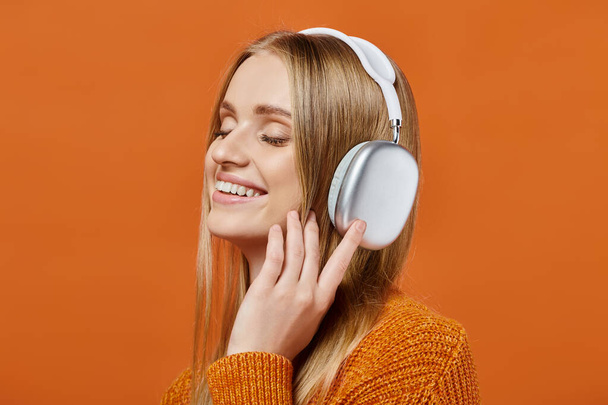 mujer rubia encantada en suéter brillante y auriculares escuchando música con los ojos cerrados en naranja - Foto, Imagen